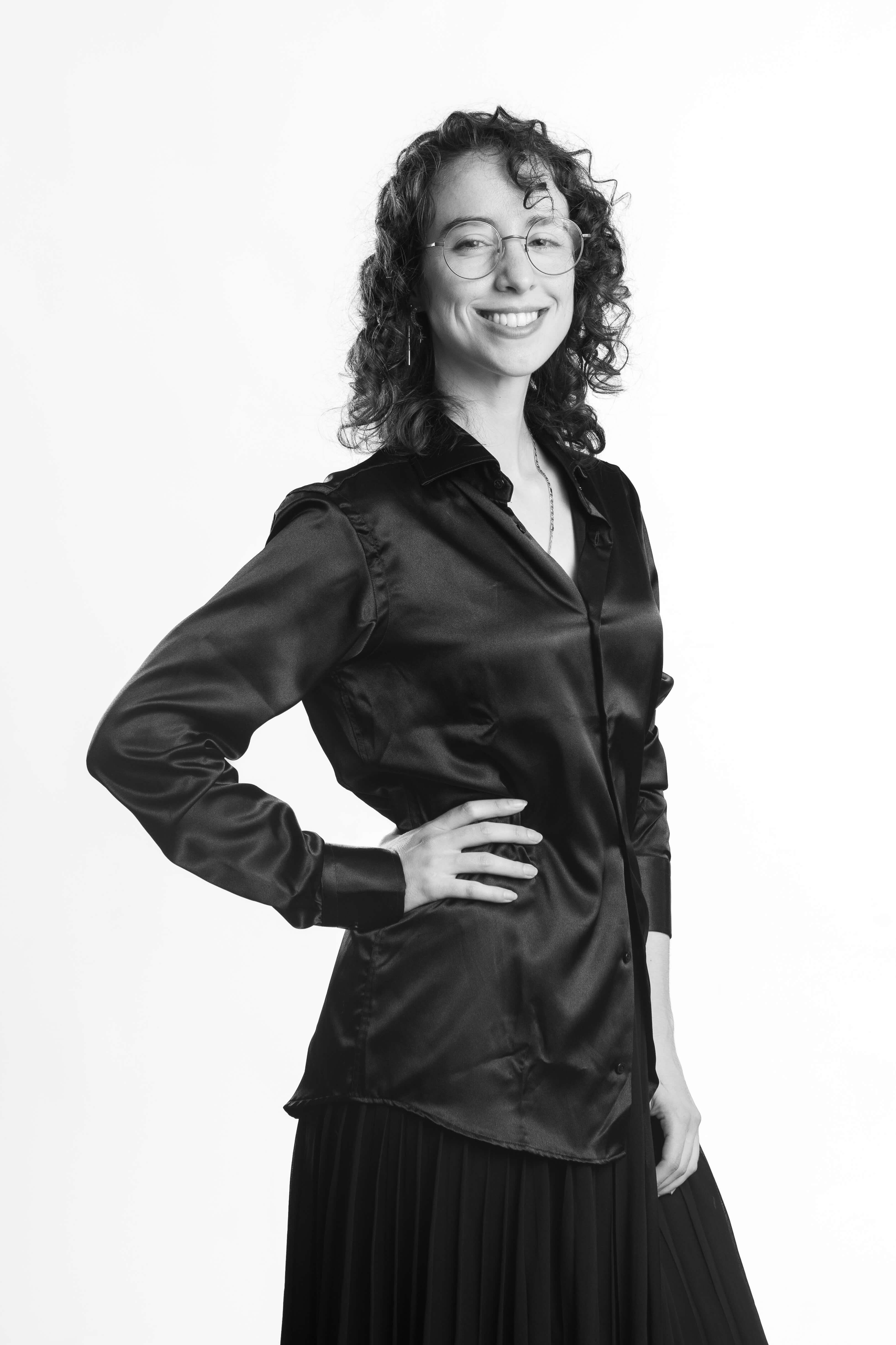 Black-and-white portrait of a smiling person with curly hair, glasses, and a silky blouse, exuding confidence and warmth against a plain backdrop.
