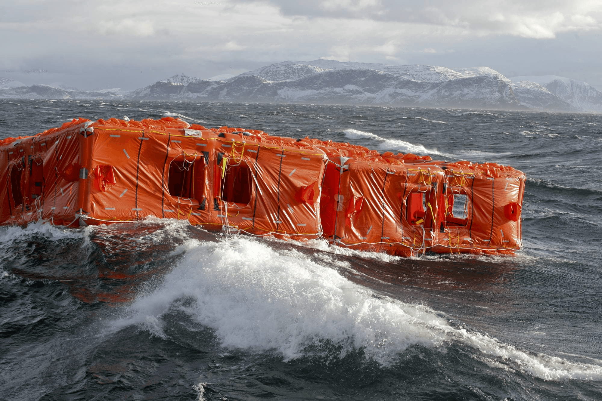 A bright orange life raft bobs on rough ocean waves under a cloudy sky, with distant land visible in the background.