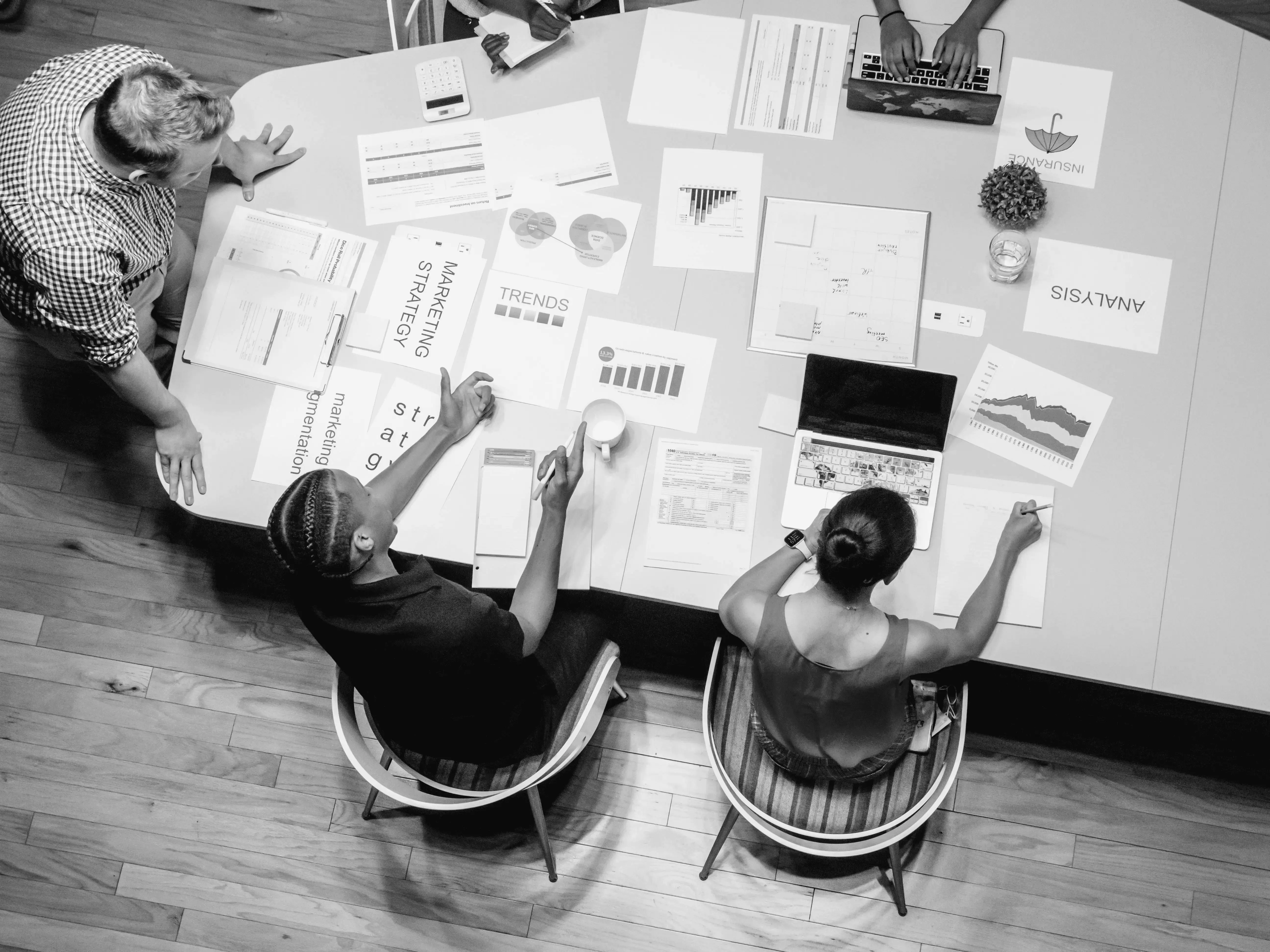 An overhead black and white view of two people at a desk covered in charts and data