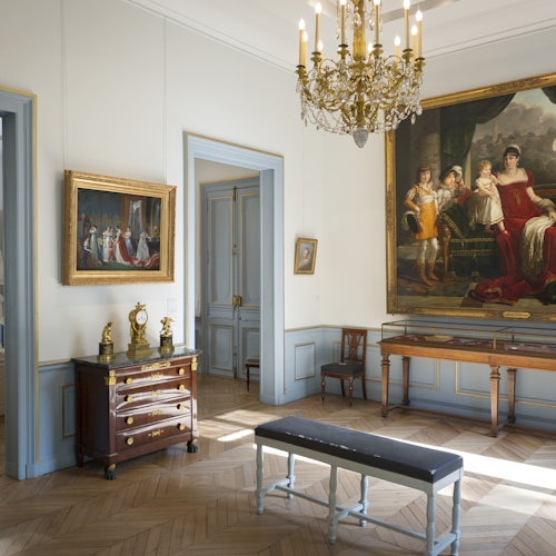 Elegant room with wooden flooring, chandelier, large paintings on the walls, antique furniture, gold accents, and a bench.