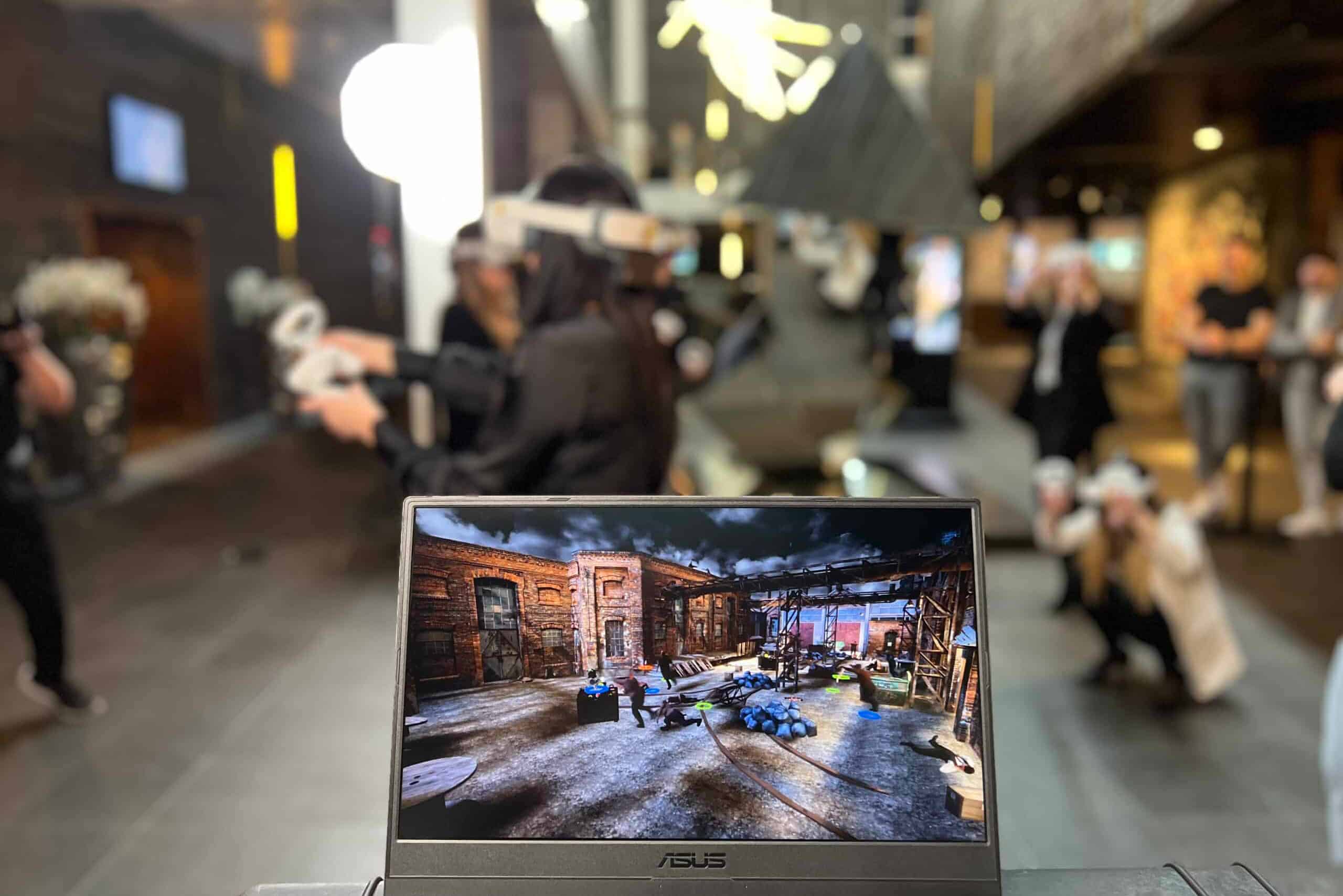 A young person with curly hair smiles at the camera while sitting in front of a laptop against a dark background.