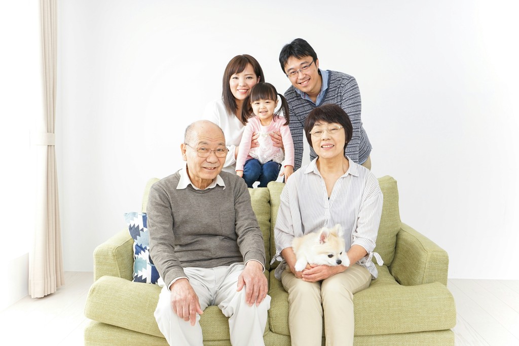 A multi-generational Asian family, including grandparents, parents, and a child, gather on a green couch in a bright, minimalist living room, with a small white dog adding a touch of warmth to the scene.