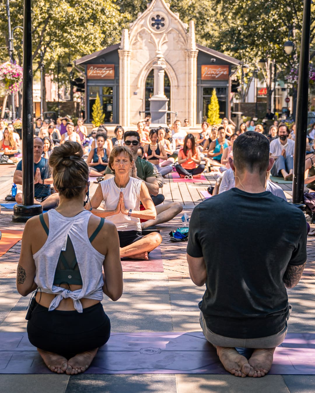 Bre and Flo leading a group yoga session outdoors, with people seated cross-legged on mats, meditating. A stone pavilion and leafy trees create a serene backdrop.