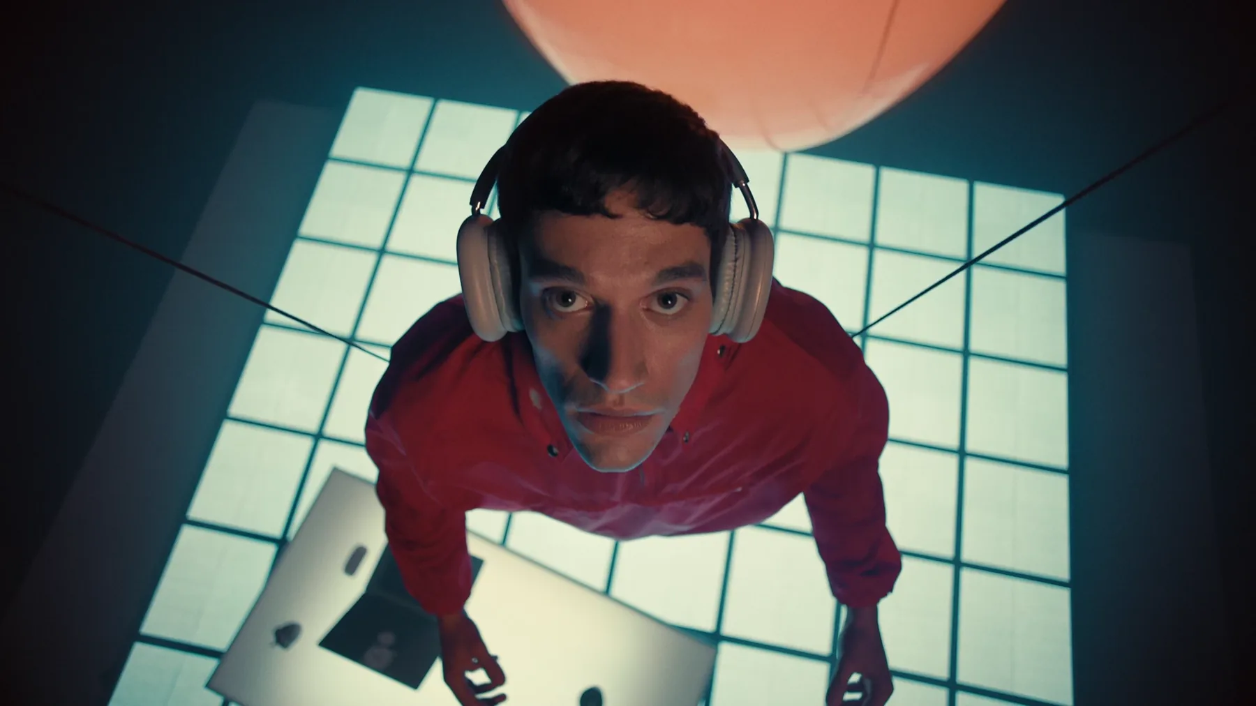 Top-down angle on a man in a red shirt looking up from a cyan illuminated grid platform, a warm orange pendant lamp glowing above him.