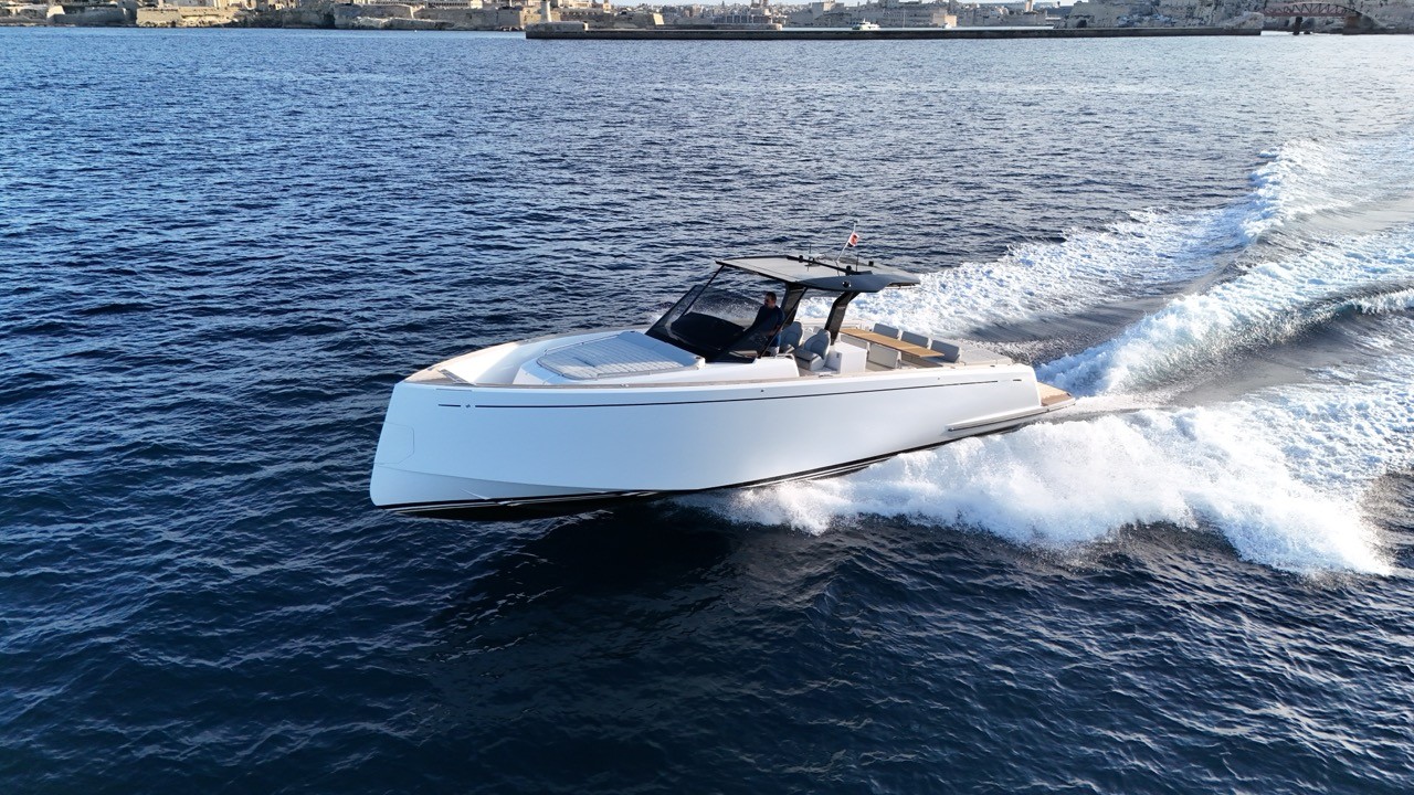 Dynamic aerial starboard three-quarter view of the Pardo 43 at high speed off Valletta, Malta, with dramatic white bow spray and wake, showing the planing hull attitude, full-length teak deck, T-top hardtop with driver at the helm, foredeck sunpad, and the Valletta Grand Harbour fortifications stretching along the horizon