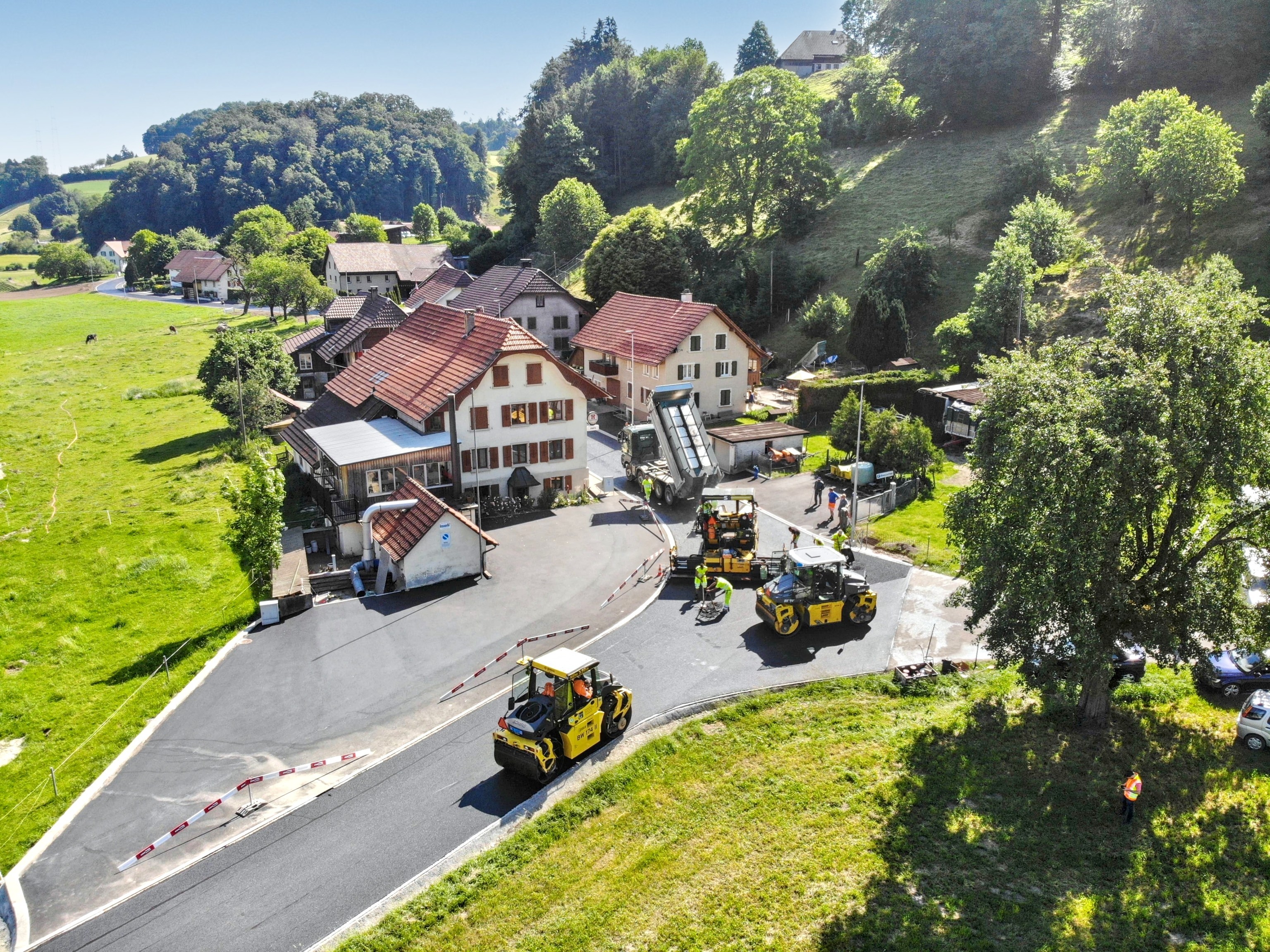 Aerial view of Strub Bau AG completing asphalt paving in a rural Swiss village – road construction integrated with scenic countryside.