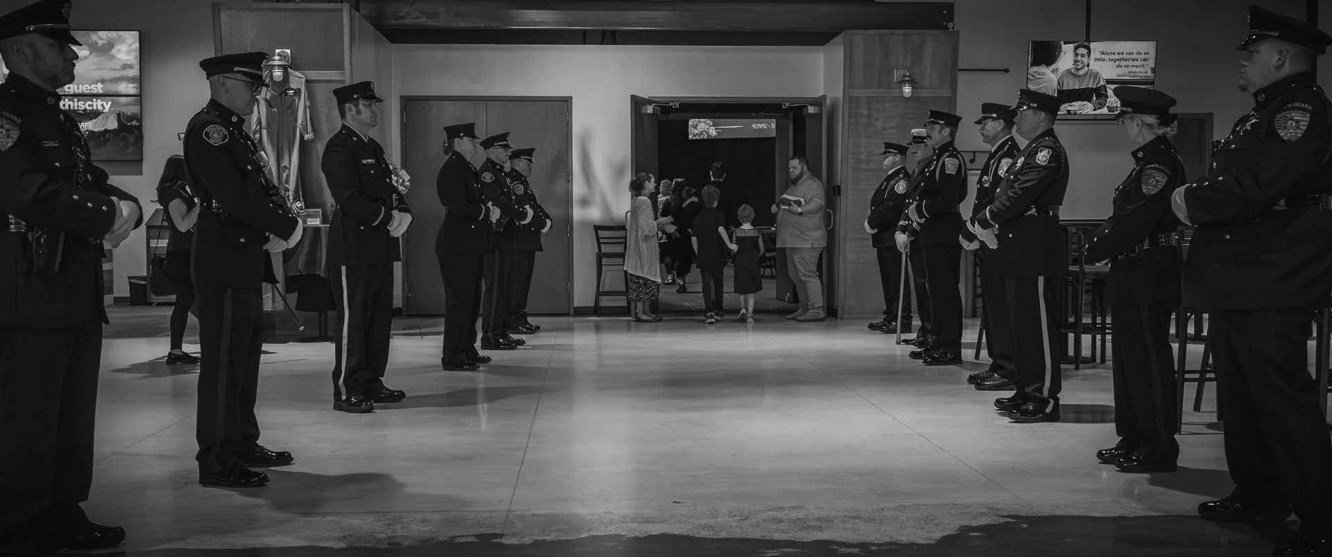 A group of uniformed officers stands attentively in two parallel lines inside a building, creating a ceremonial passageway, with people entering through the doorway in the background.