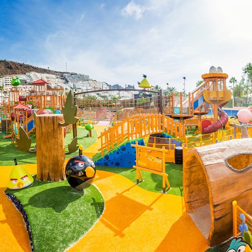 一个色彩斑斓的游乐场在阳光明媚的一天，设有滑梯、桥梁、攀爬结构和角色主题设计。背景中可以看到公寓楼。