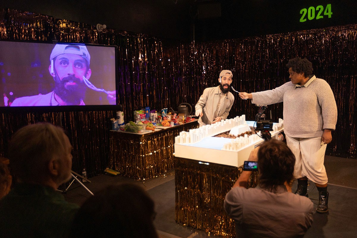 A performer acts as if he is being interviewed. His face is also shown on the projector screen.