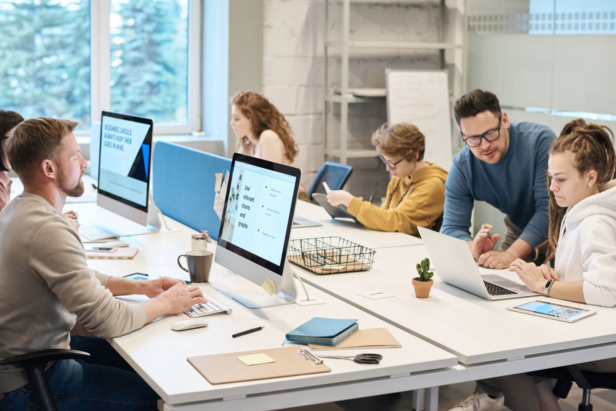 A bright, sunlit coworking space where a woman works on a desktop computer in the foreground, while another colleague in a yellow jacket works further down the long white desk organized with notebooks and small plants.