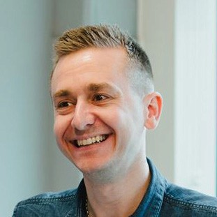 A woman with short curly hair wearing a black t-shirt looks into the camera.