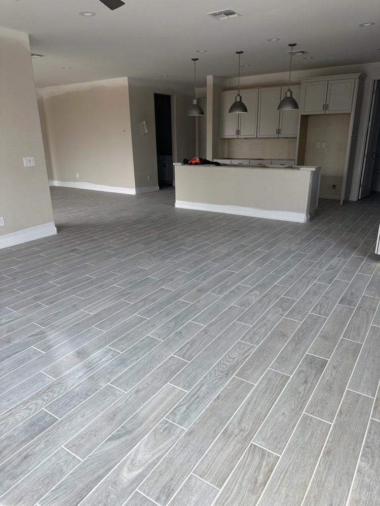 Modern open-plan living area with gray wood-look tile flooring, kitchen island, and large windows in a new home under construction.