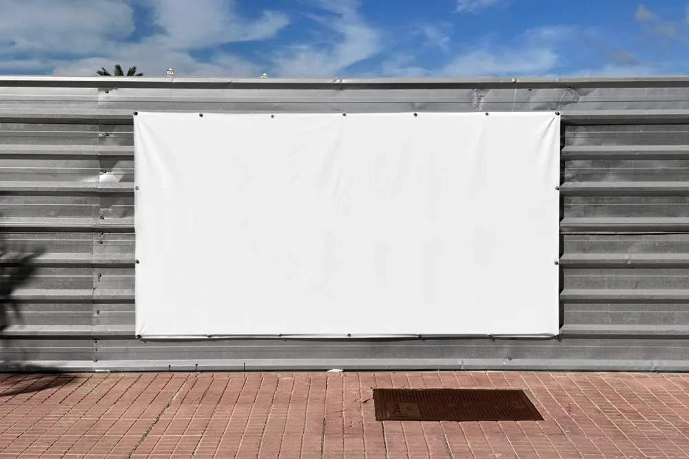 Mockup of a green banner attached to a corrugated metal fence, set against a sunny outdoor backdrop with a blue sky. The clean design and bold typography make it ideal for showcasing promotional content.