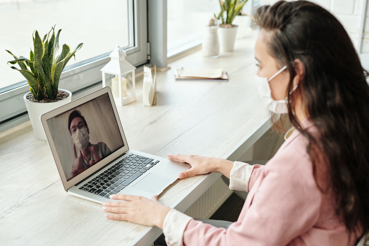 A woman sits in front of a laptop computer and video chats with a man; both are wearing face masks. Technology in Interior Design.