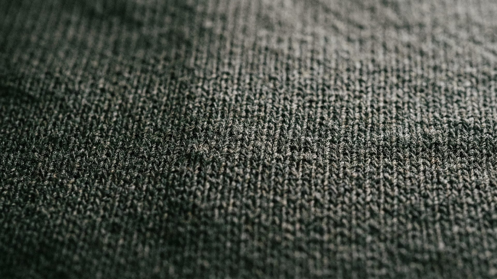 An extreme macro close-up of a premium 26 double ring-spun charcoal cotton jersey fabric, showing the tight, dense weave structure under raking overhead light.
