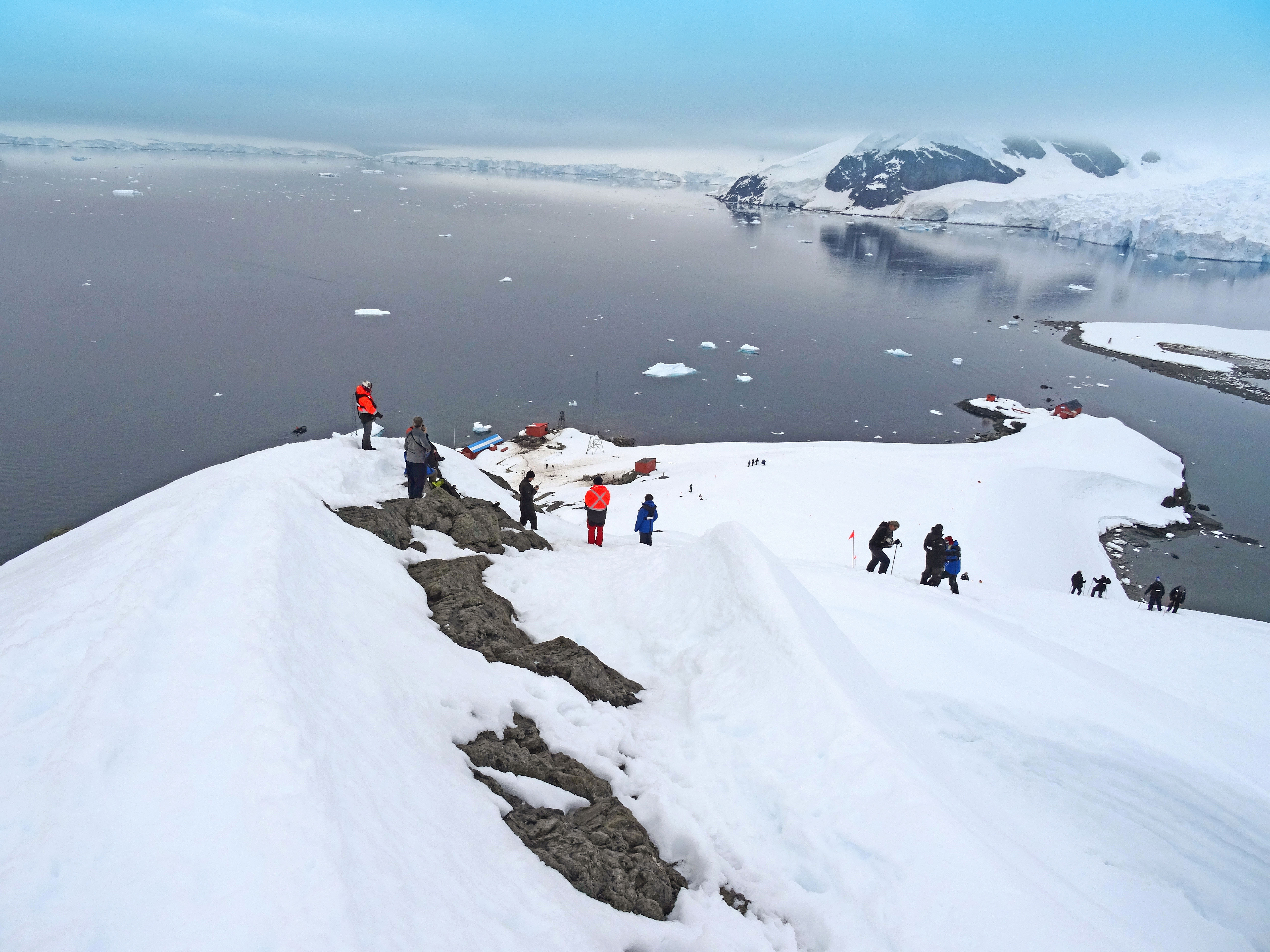 The photograph depicts a snowy landscape with a group of people hiking along a snow-covered ridge near Brown Station in Antarctica