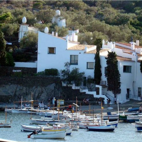 Small boats moored on water in front of white buildings built into a hillside with trees and greenery.