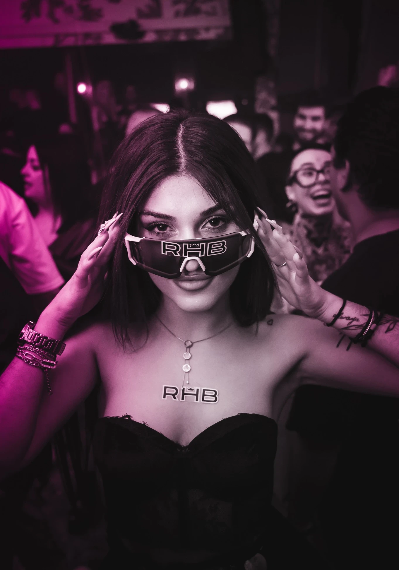 A woman in a black strapless top, an "RHB" necklace, and an "RHB" sticker on her chest looks at the camera while adjusting a pair of large, futuristic sunglasses also branded with "RHB".