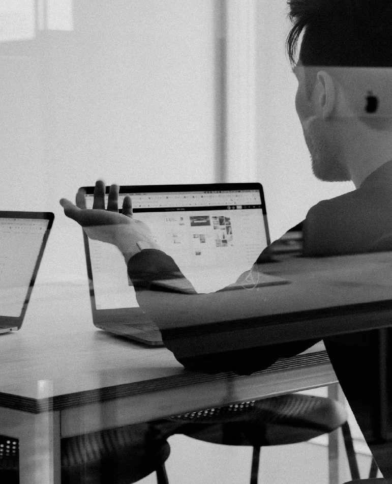 A black and white image of a guy talking to someone sitting with his back towards the camera and an open laptop in front of him