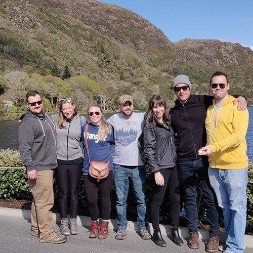 Шест души стоят близо един до друг и се усмихват на камерата. Те се намират на открито с планини и езеро на заден план.