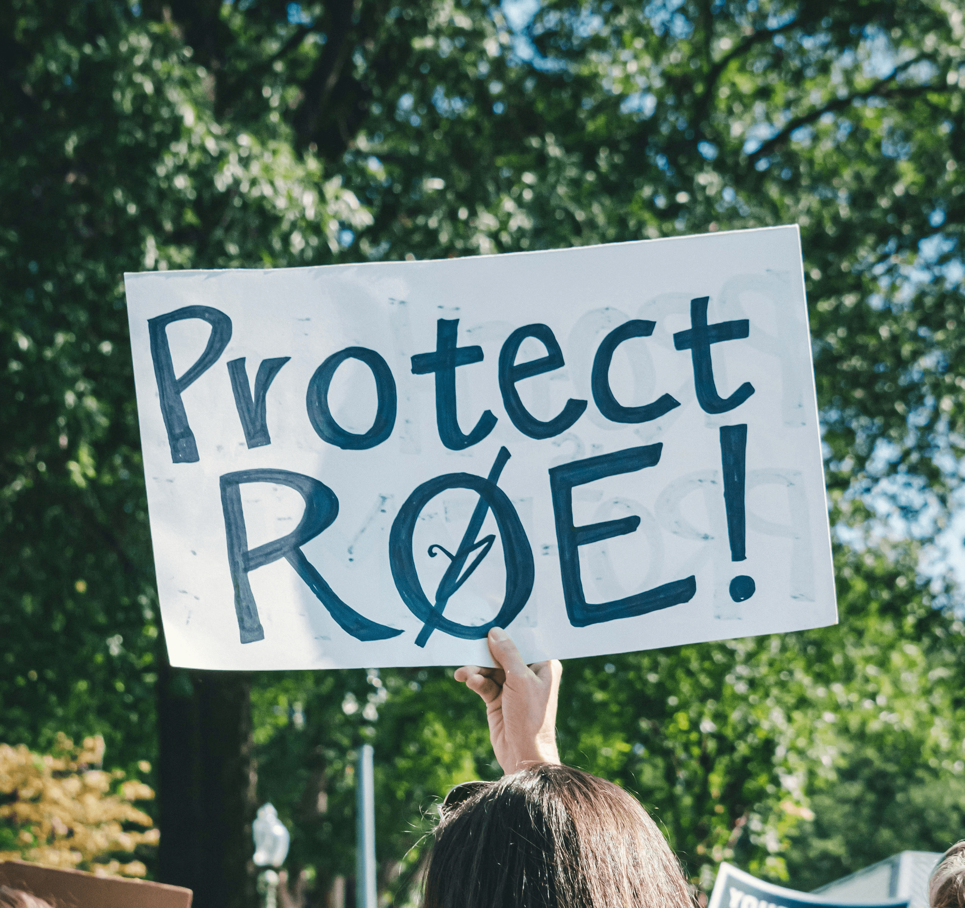 A protestor holding up a sign with one hand that reads, "Protect Roe" and has a slash through the "o" with the slash also going through a coat hanger