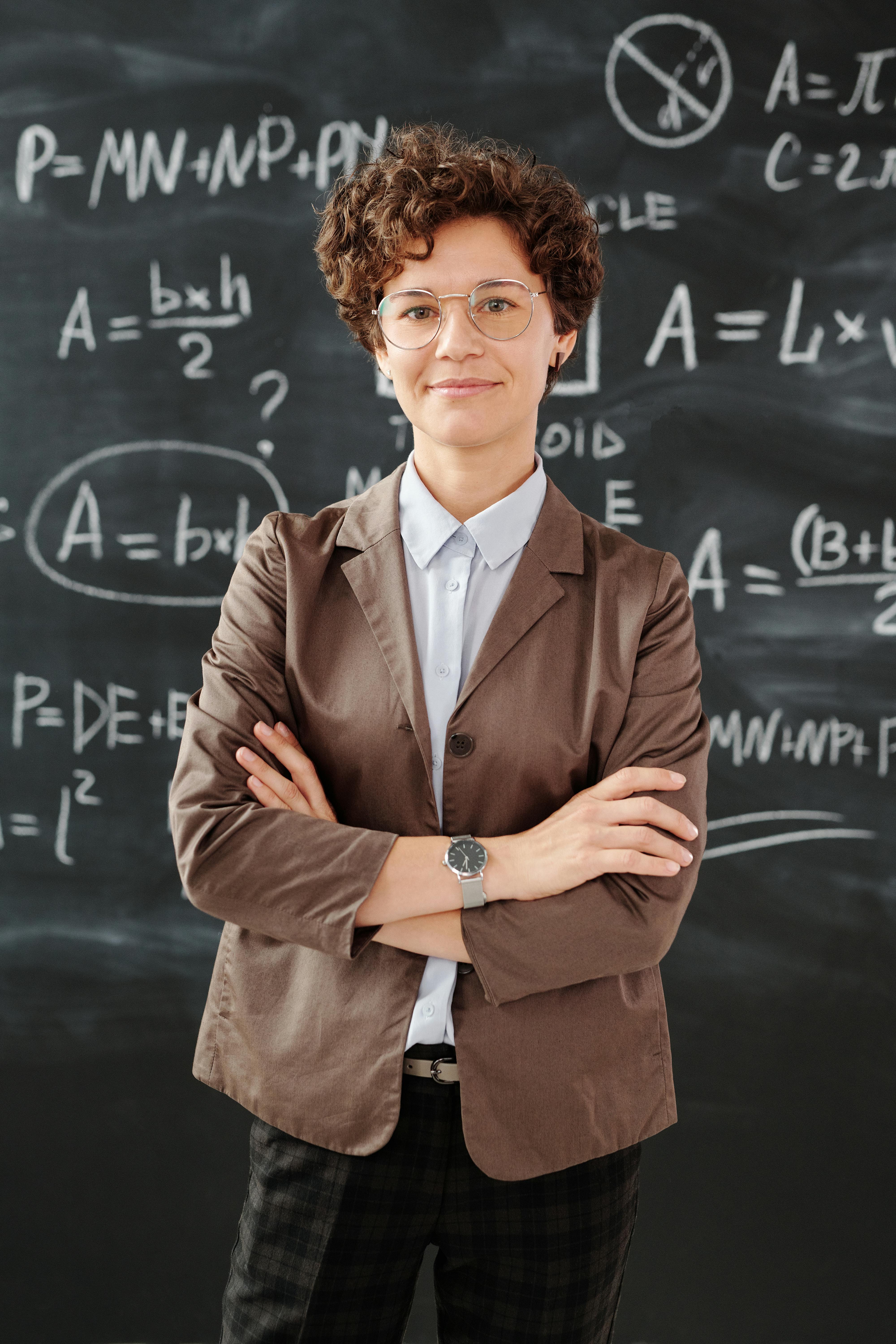 Woman in glasses and blazer in front of chalkboard.