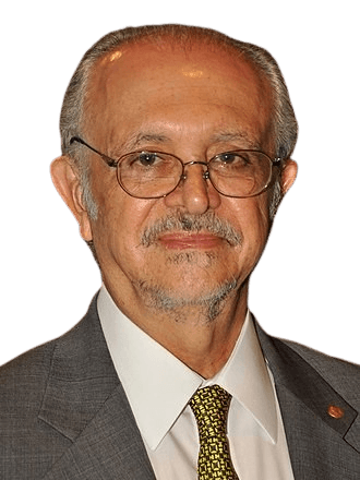 A color photograph of an older man with glasses, a neatly groomed mustache, and a receding hairline, wearing a gray suit, white shirt, and yellow patterned tie. He has a calm, composed expression, and the warm indoor lighting suggests a formal or professional event. The background is softly blurred, keeping the focus on his distinguished appearance.