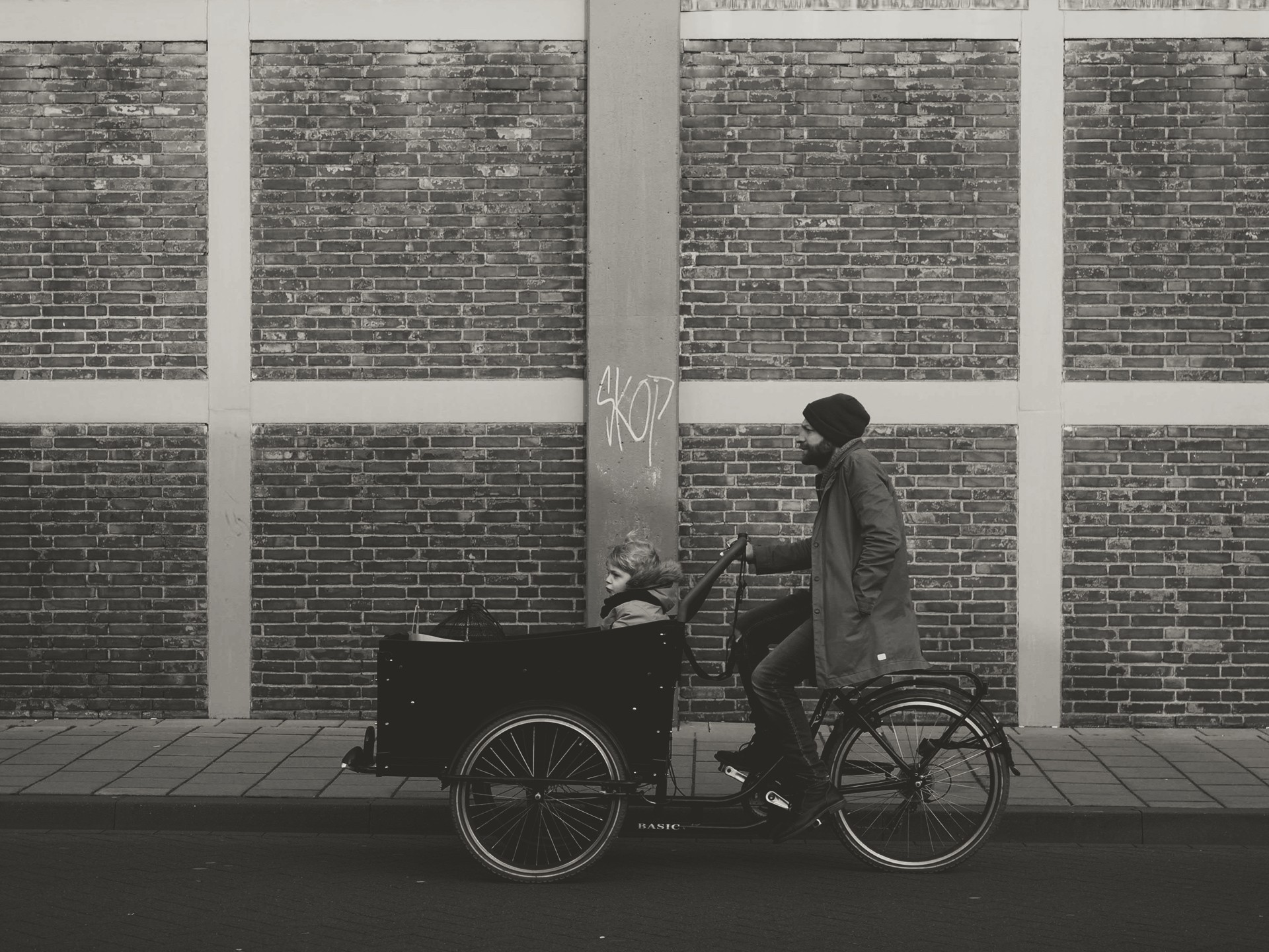 Urban black-and-white scene showing a cargo bicycle carrying a child on a sidewalk next to a brick-and-concrete building, evoking everyday city life and movement.