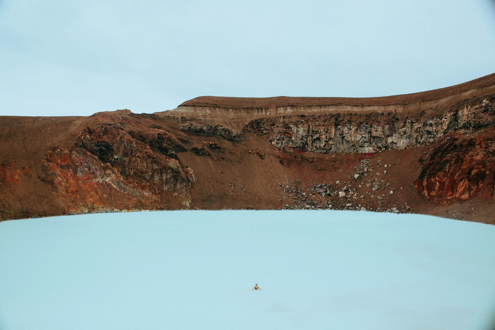 A pale green crater lake ringed by red and grey volcanic walls