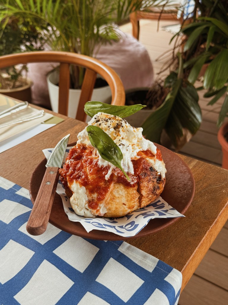A plate featuring a large bread roll topped with marinara sauce, fresh cream, and a basil leaf, on a wooden table with lush green plants in the background.