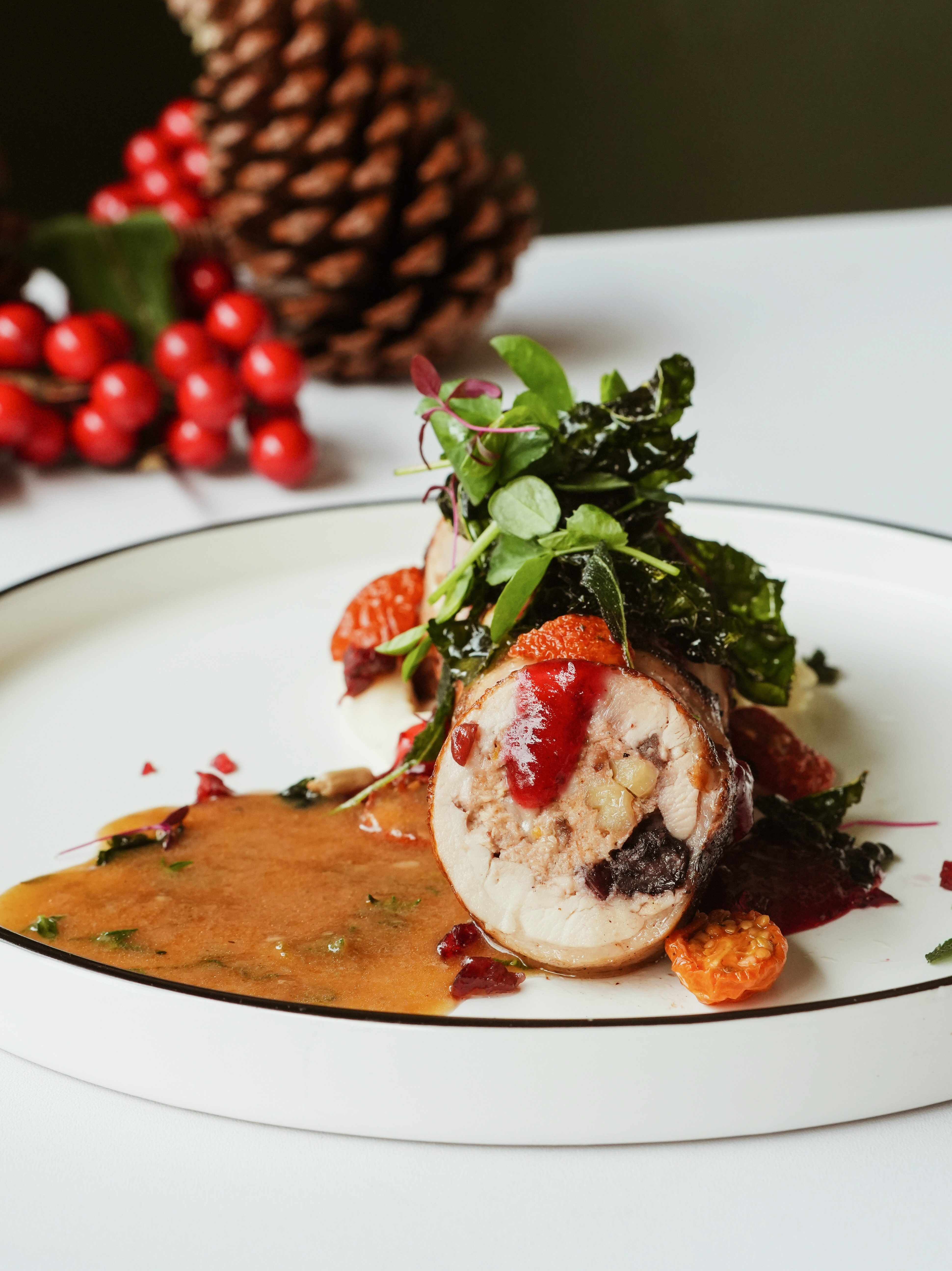 A beautifully plated Christmas dish featuring rolled meat with garnishes, accompanied by a savory sauce, set against a festive backdrop of pinecones and holiday decor.