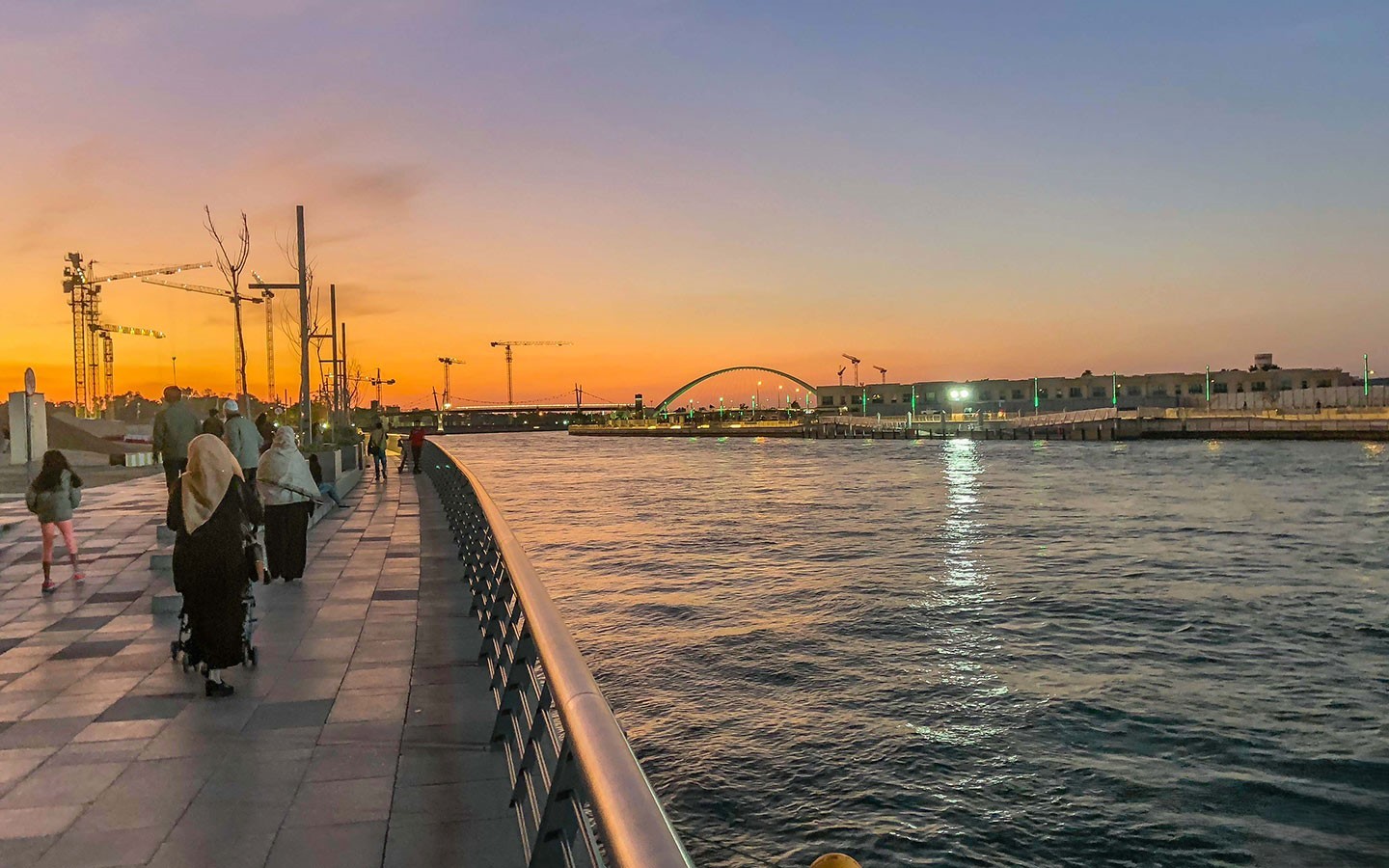 People walk along the waterfront promenade of Dubai Design District (d3) at sunset, with a bridge arching in the distance.