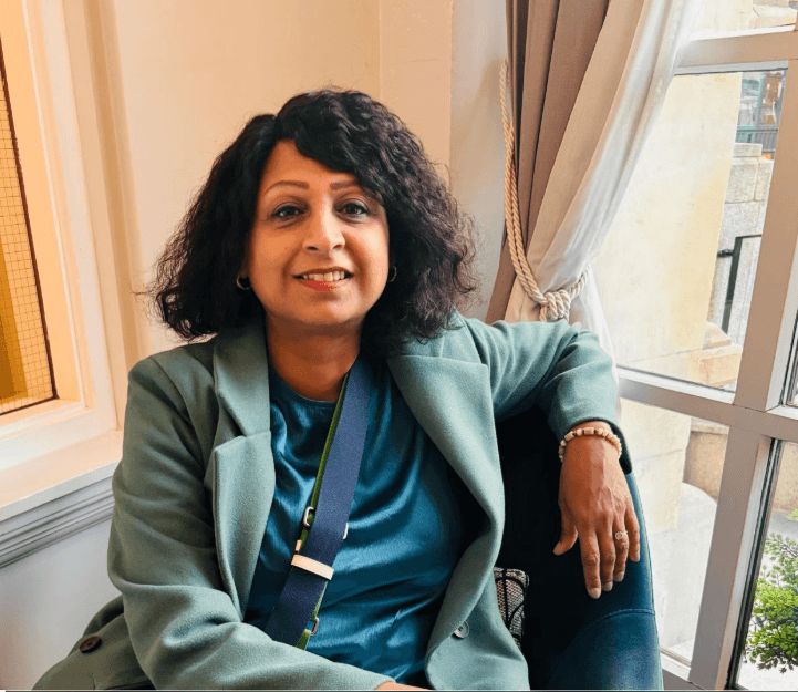 Close-up portrait of a blonde woman in translucent pink glasses and a white shirt on a soft blue background.