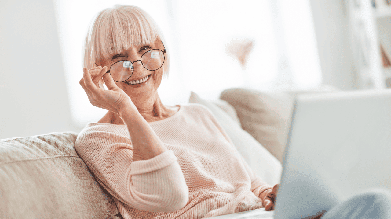 Old woman smiling in front of her laptop while holding her glasses