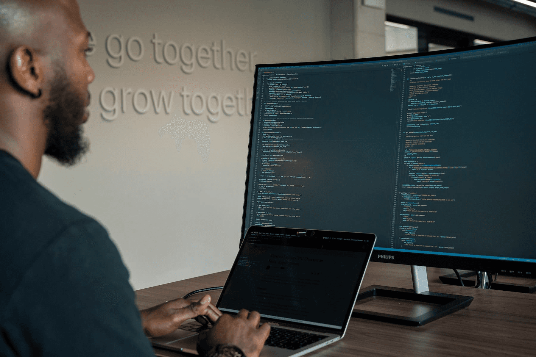 A person focuses on writing and reviewing code displayed on a laptop and a large monitor in a modern office, emphasizing technology and software development teamwork.
