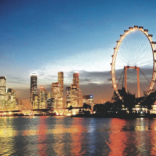 City skyline at dusk with illuminated skyscrapers and a large Ferris wheel by the water, reflecting colorful lights.