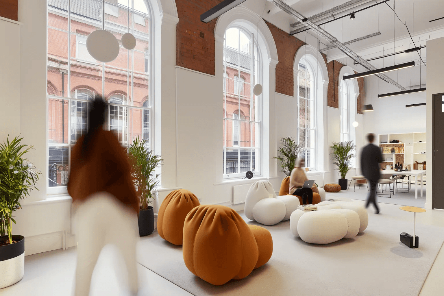 Modern open office lounge with large arched windows, plants, and soft round seating in white and terracotta tones, with people walking and working on laptops.