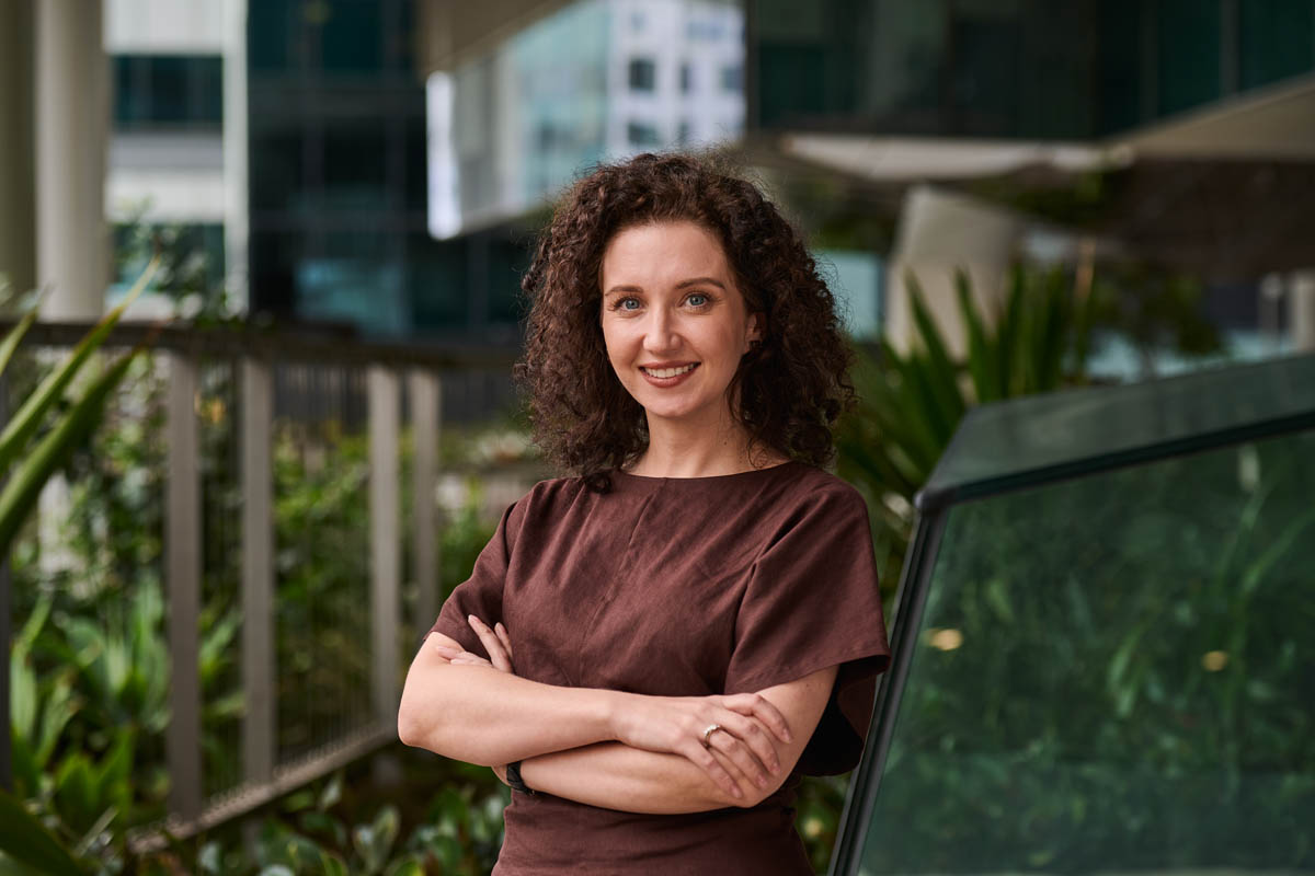 Corporate headshot of woman with curly hair in brown top, outdoor office background