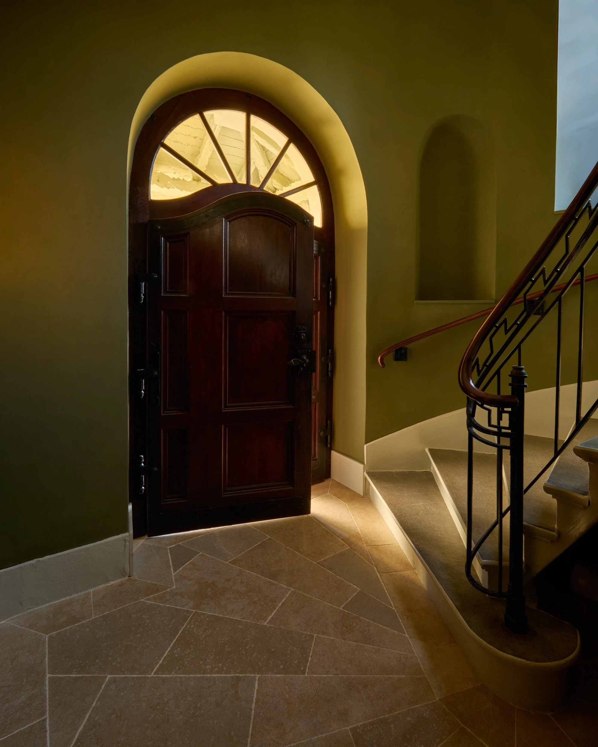 Entrée de la cage d’escalier du XIXᵉ siècle au Château de Saint-Prex, avec porte principale en chêne, début de l’escalier en spirale et sol en pierre calcaire beige.