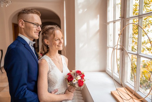 Brautpaar steht am Fenster und blickt ins Licht während Hochzeit im Innenraum