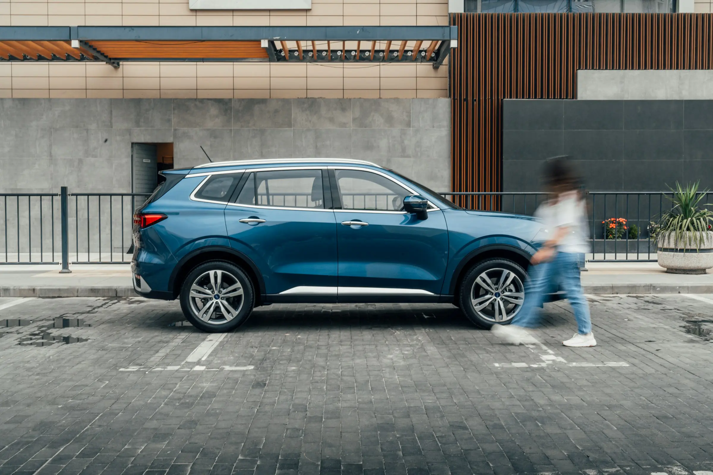Blue SUV parked on a city street in front of a modern building, with a person walking past in motion blur.