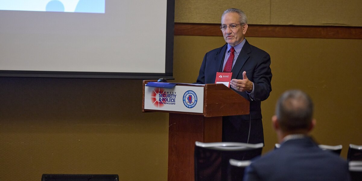 An individual stands at a podium giving a presentation at a conference, looking confident and engaged. A projector screen is partially visible in the background.