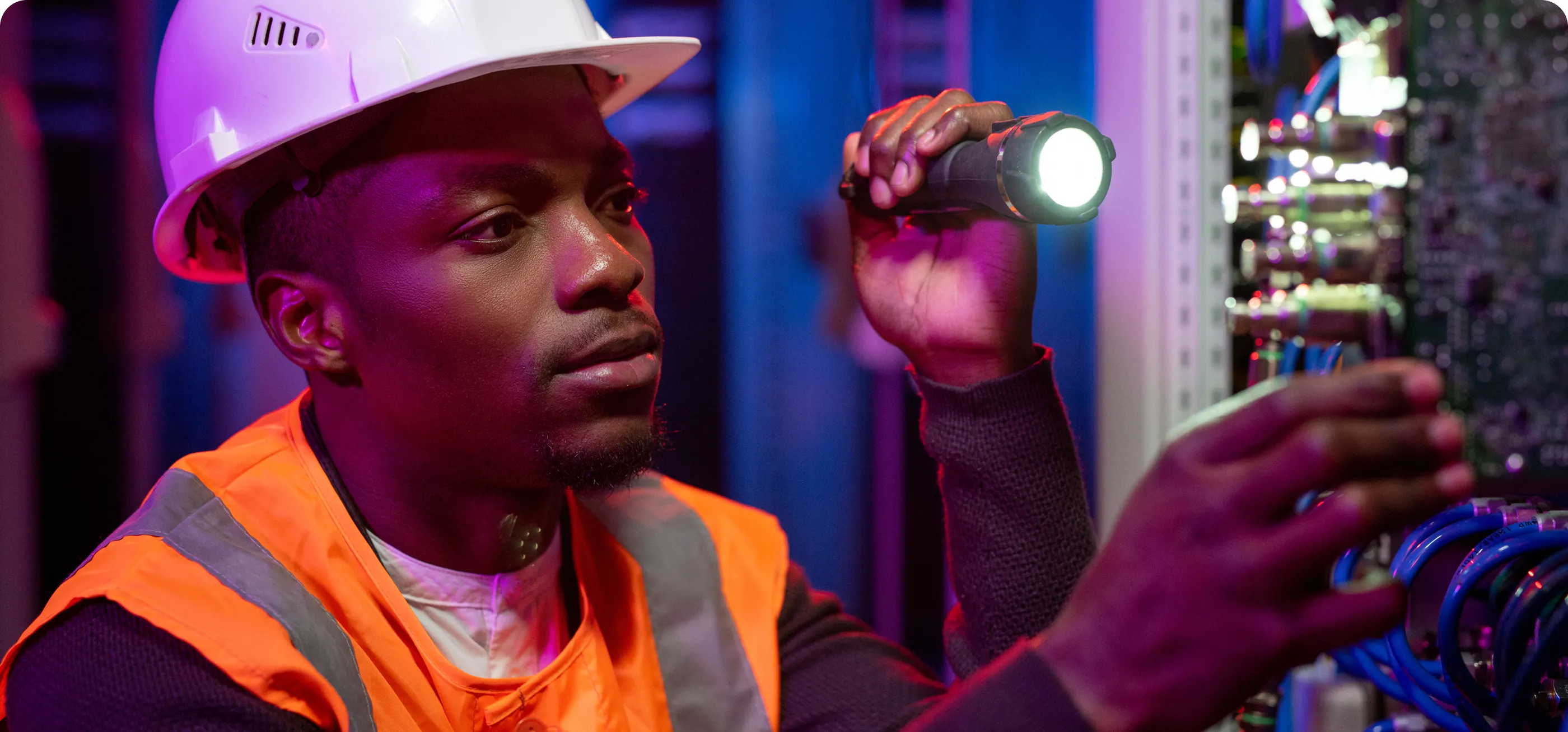 Technicien en gilet orange inspecte un panneau électrique avec une lampe.