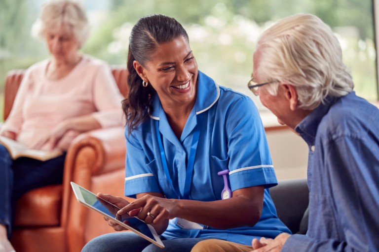 A care worker in blue nursing uniform sitting beside an older woman in a care home setting, engaged in personal care