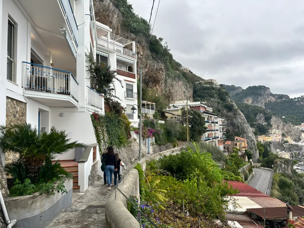 A narrow stepped alley in Amalfi between white houses, with two people walking down towards the sea.