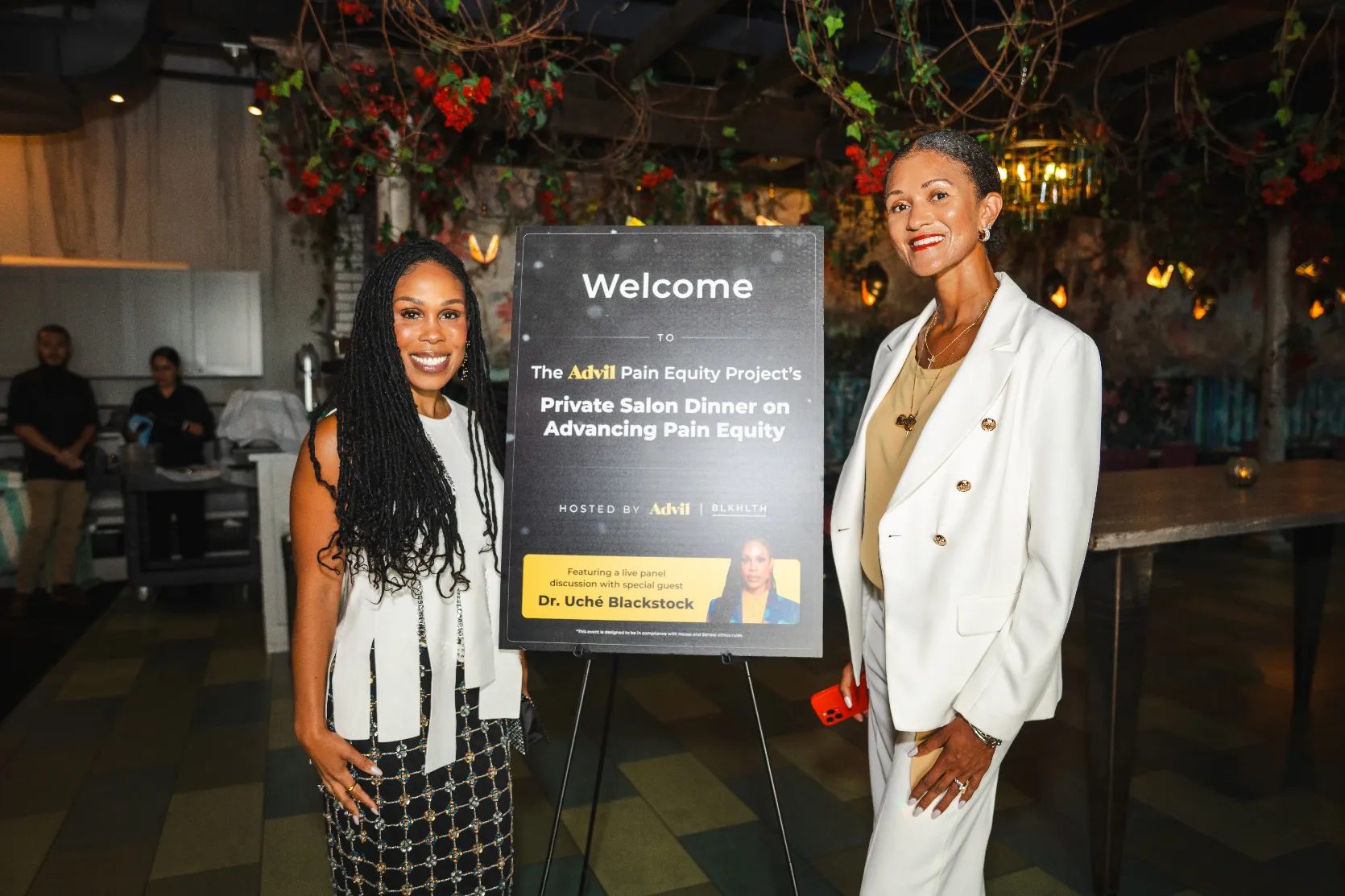 Two women smile beside a sign that reads “Welcome. The Advil Pain Equity Project’s Private Salon Dinner on Advancing Pain Equity,” featuring Dr. Uché Blackstock. The setting is elegant with plants and warm lighting.