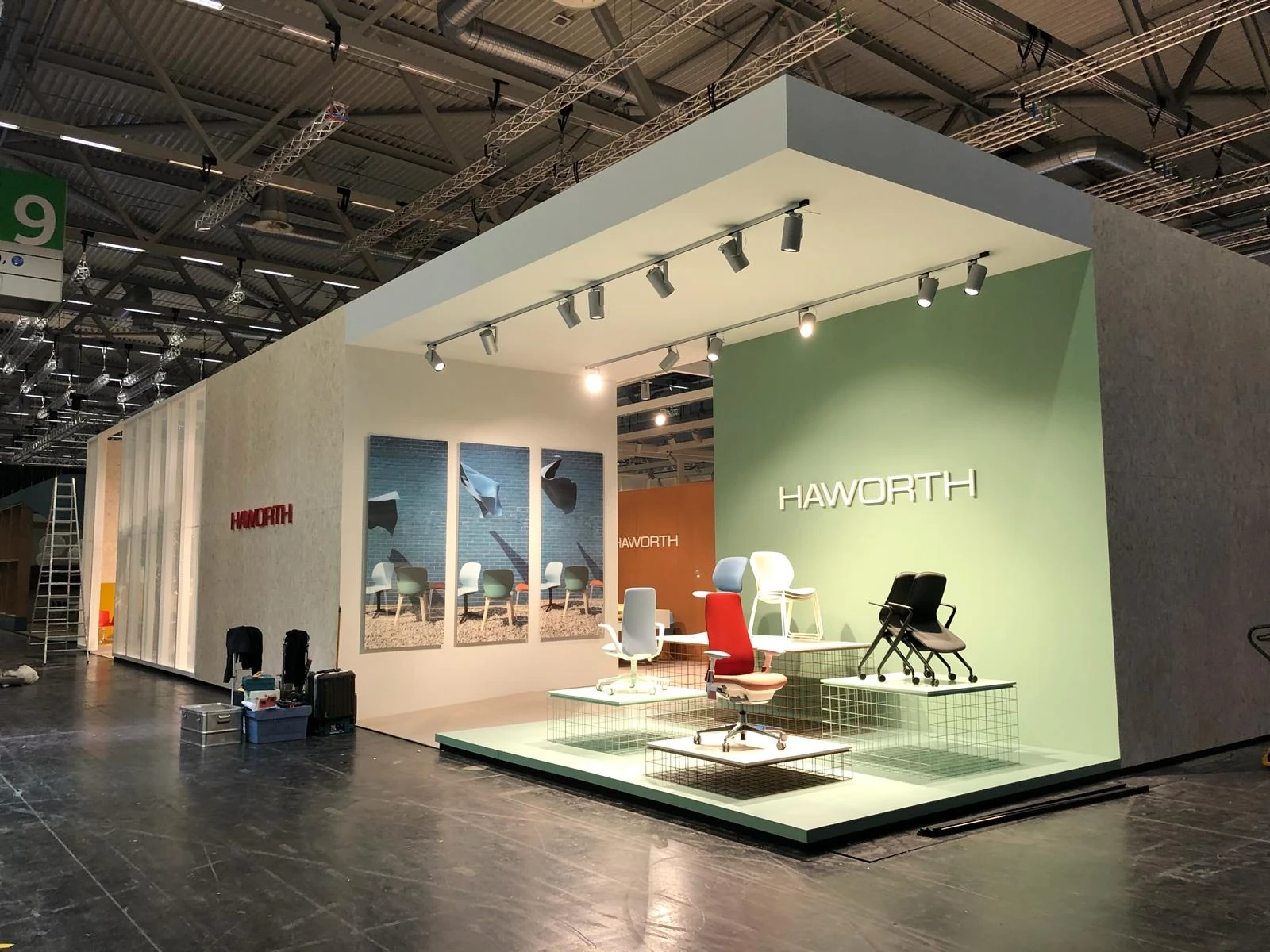 Overall view of the HAWORTH ORGATEC trade fair stand with office chairs on white podests in front of a sage-green wall with white logo.