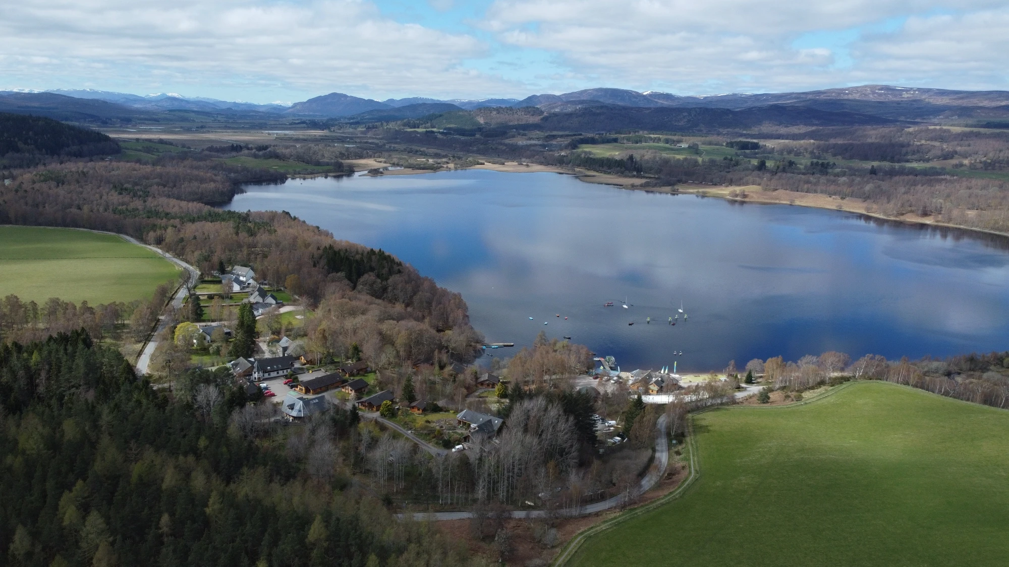 Aerial view of a lochside area with a sandy beach, surrounded by forest and mountains. Buildings and a dock with boats are visible, under a clear blue sky.