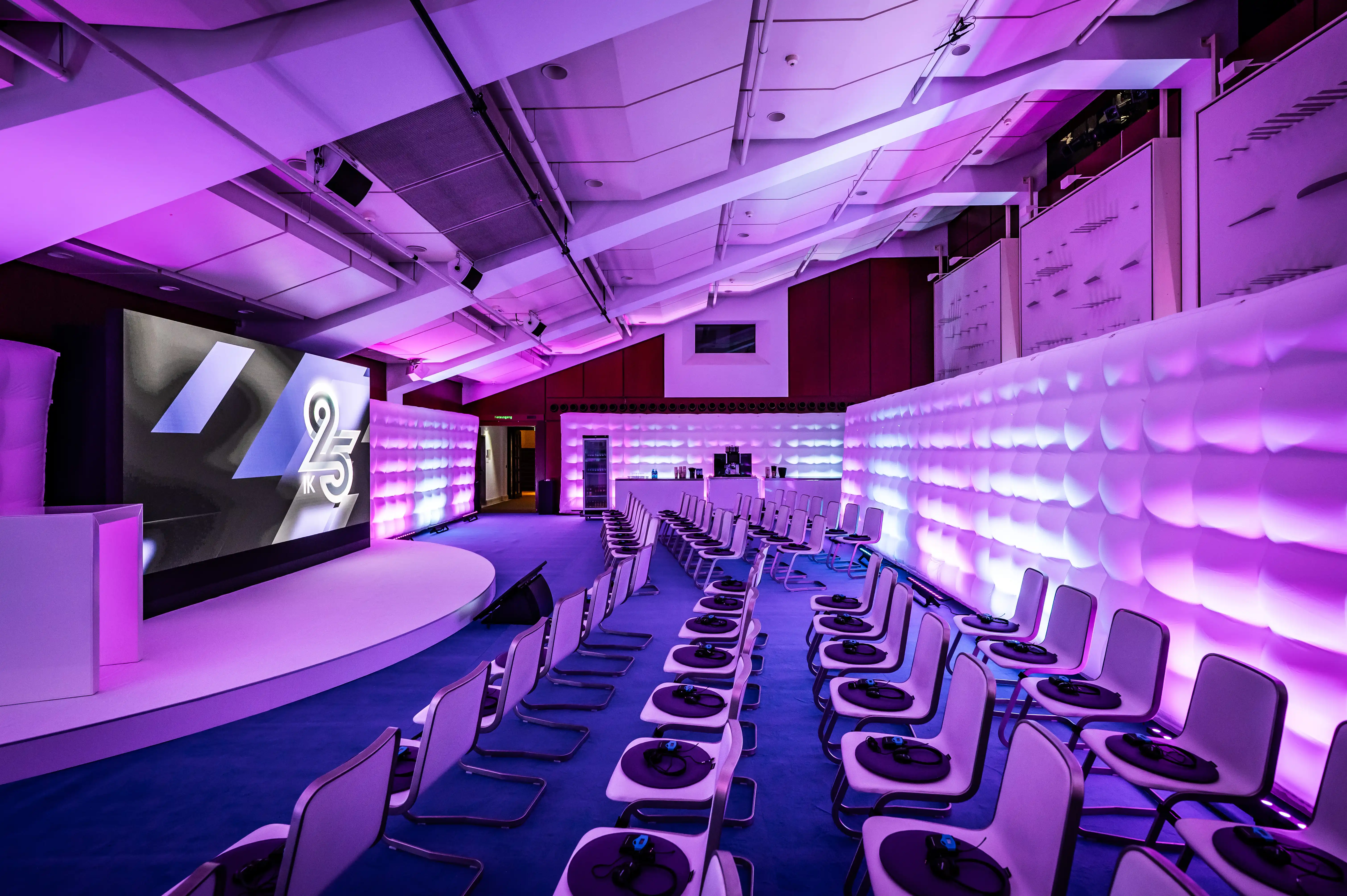 Conference room in purple light with white quilted walls, stage and rows of chairs with desks.