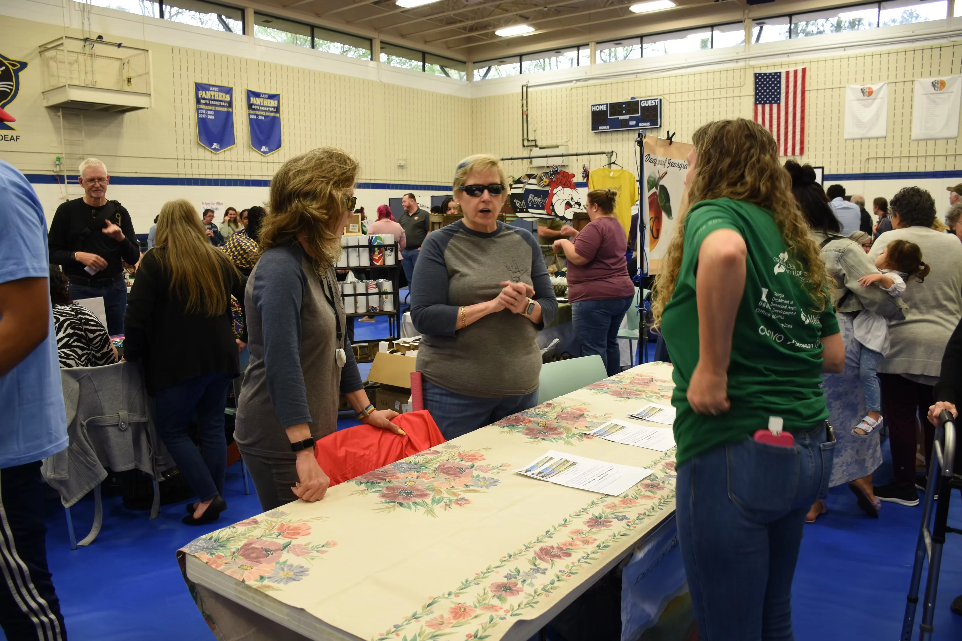 People gather at a table in a gymnasium, browsing and chatting at a community event or fair.